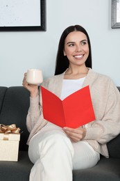Happy woman reading greeting card while drinking coffee on sofa in living room