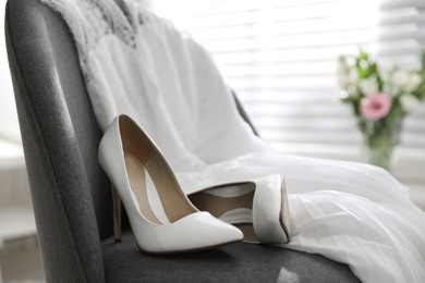Photo of Pair of white high heel shoes and wedding dress on chair indoors