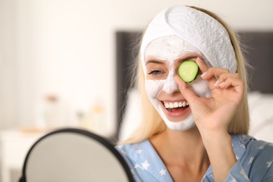 Photo of Young woman with face mask and cucumber looking into mirror at home, space for text. Spa treatments