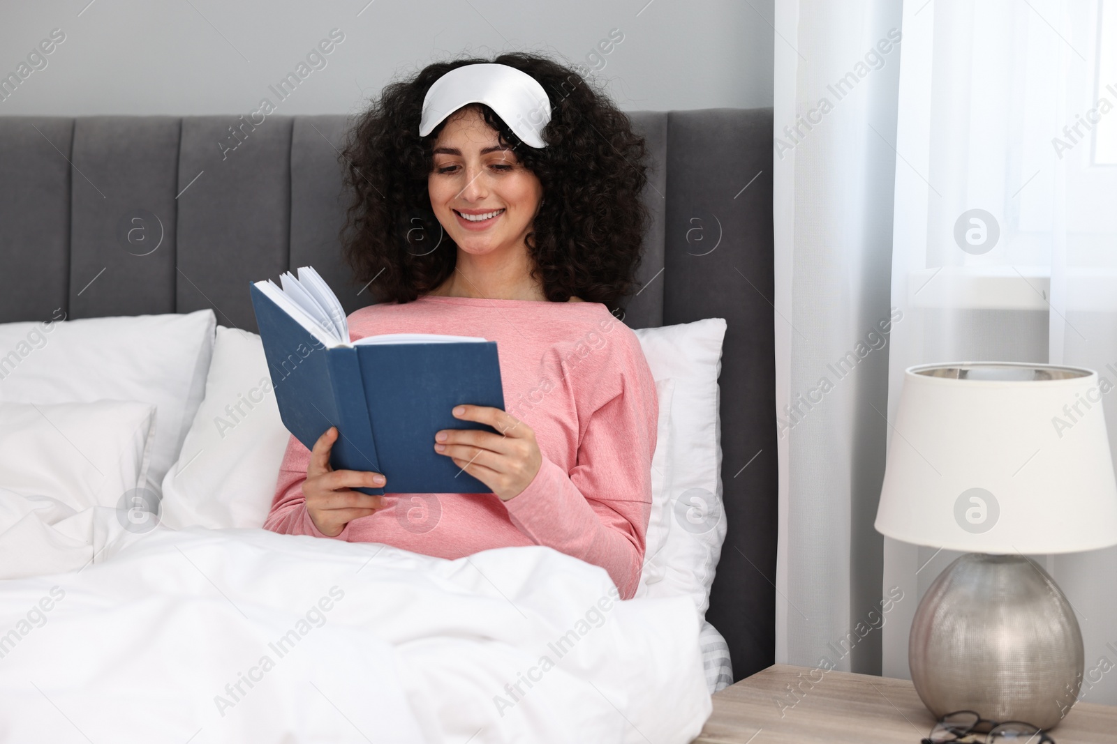 Photo of Beautiful young woman in stylish pyjama and sleep mask reading book on bed at home