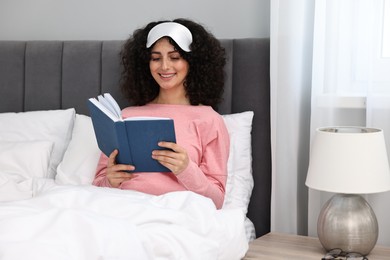 Photo of Beautiful young woman in stylish pyjama and sleep mask reading book on bed at home