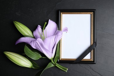 Image of Funeral photo frame with ribbon and violet lily on black table, flat lay