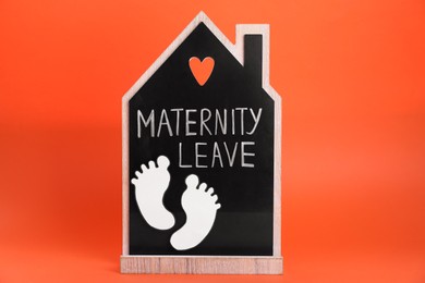 Photo of Wooden house figure with words Maternity Leave and paper cutout of baby feet on orange background