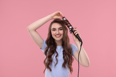 Photo of Beautiful young woman using curling hair iron on pink background