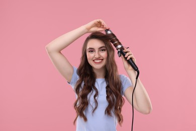 Beautiful young woman using curling hair iron on pink background