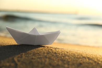 Photo of White paper boat on sand near sea at sunset, closeup. Space for text