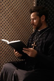 Catholic priest in cassock reading Bible in confessional booth