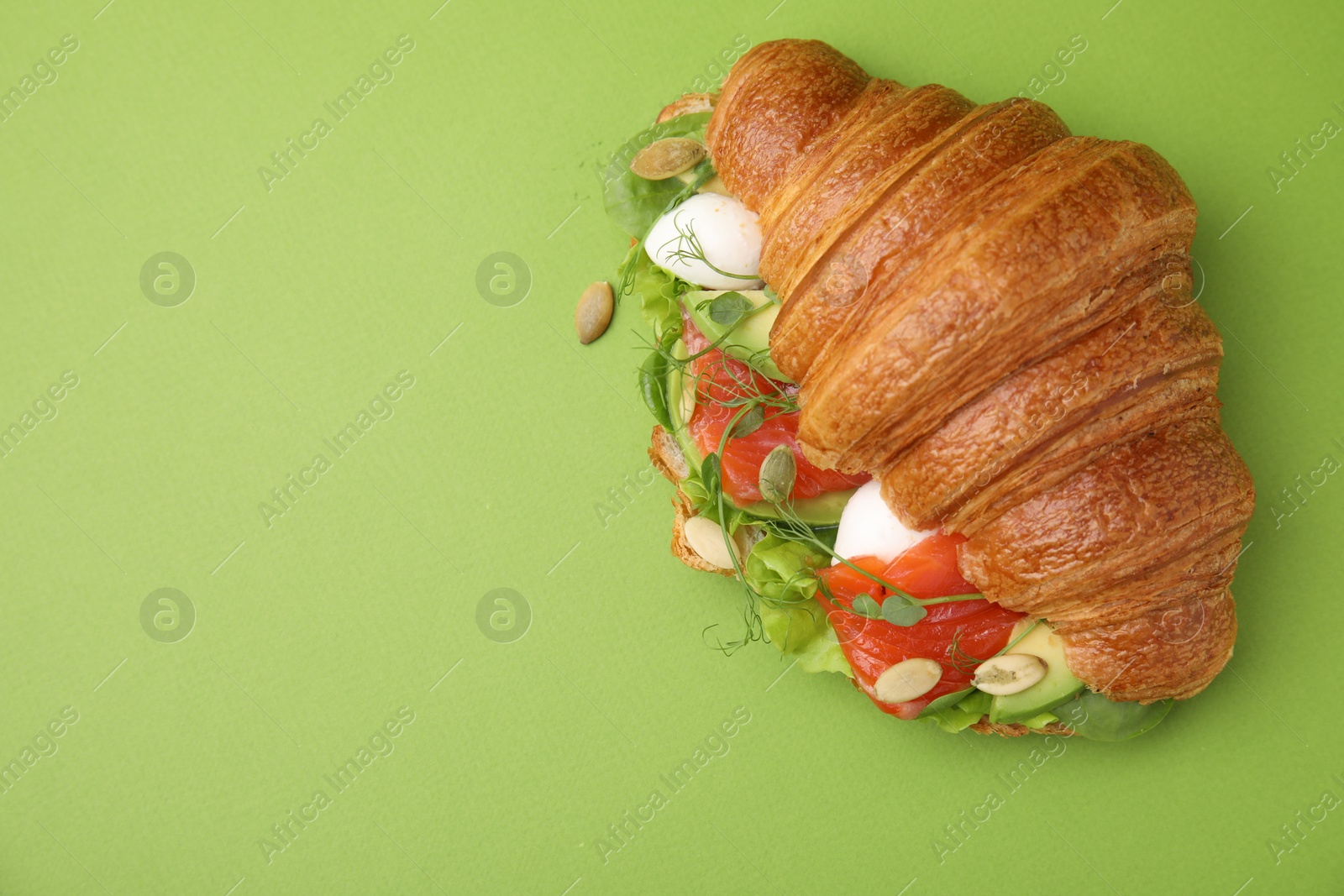 Photo of Tasty croissant with salmon, avocado, mozzarella and lettuce on green background, top view. Space for text