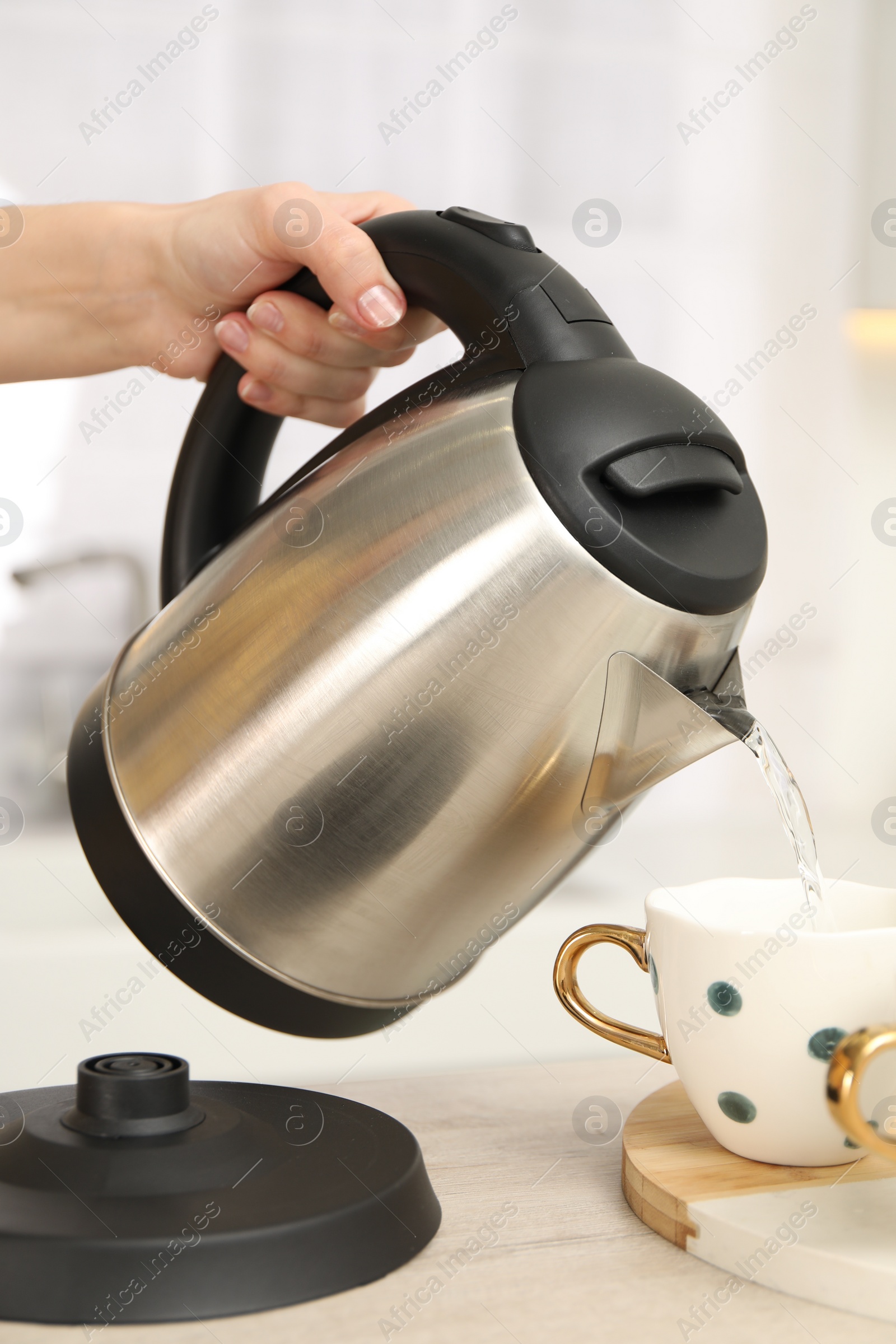 Photo of Woman pouring hot water from electric kettle into cup in kitchen, closeup