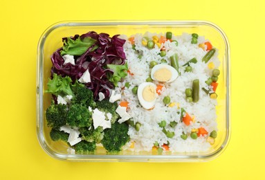Photo of Tasty rice with vegetables and salad in glass container on yellow background, top view