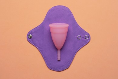 Menstrual cup and cloth pad on pale orange background, top view. Reusable feminine hygiene products