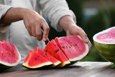 Photo of Man cutting tasty ripe watermelon at wooden table outdoors, closeup