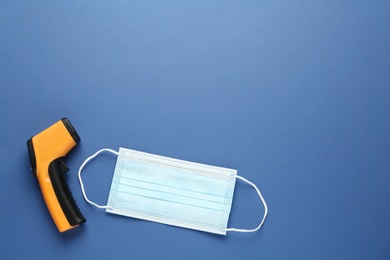 Infrared thermometer and medical mask on blue background, flat lay with space for text. Checking temperature during Covid-19 pandemic