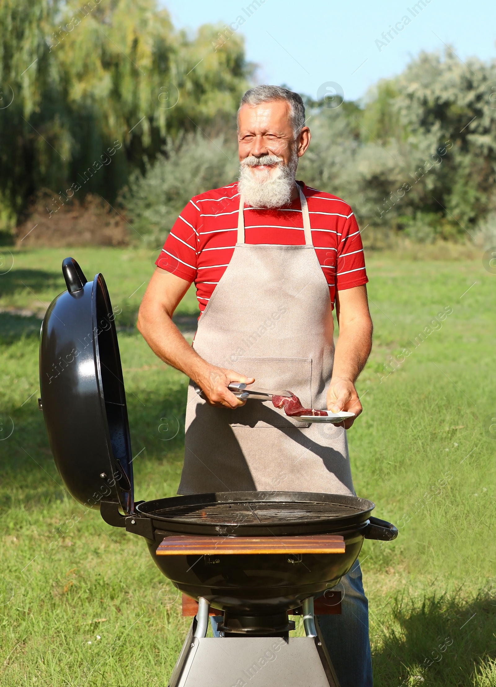 Photo of Happy senior man with meat at barbecue grill in park