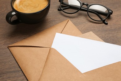 Photo of Envelopes with blank paper card, cup of coffee and glasses on wooden table, closeup