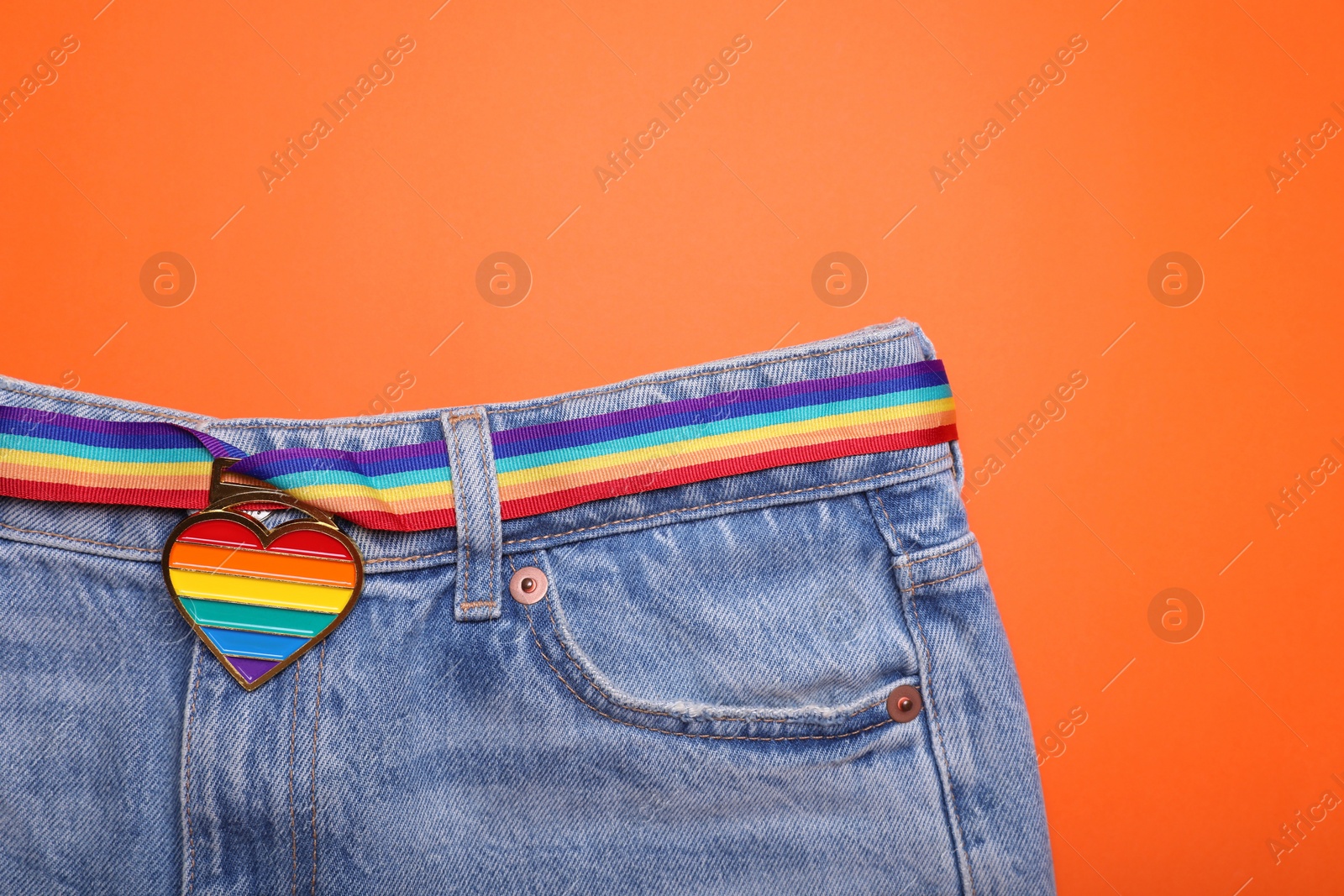 Photo of Jeans and rainbow ribbon with heart shaped pendant on orange background, top view and space for text. LGBT pride