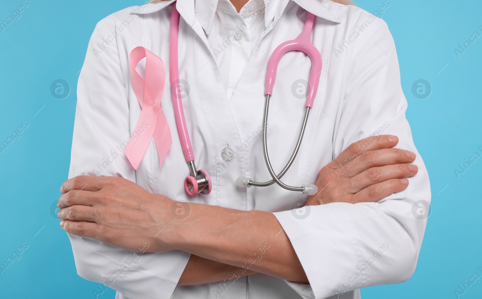 Photo of Doctor with pink ribbon and stethoscope on light blue background, closeup. Breast cancer awareness