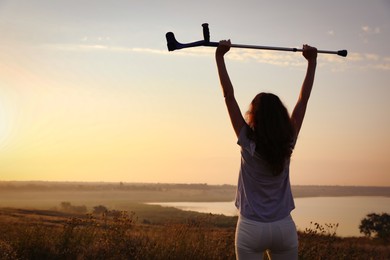 Photo of Woman raising hands with elbow crutch up to sky outdoors at sunrise, back view. Healing miracle