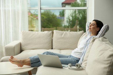 Young woman with laptop and headphones relaxing on sofa at home, space for text
