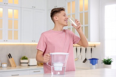 Photo of Happy man drinking clear water near filter jug at table in kitchen