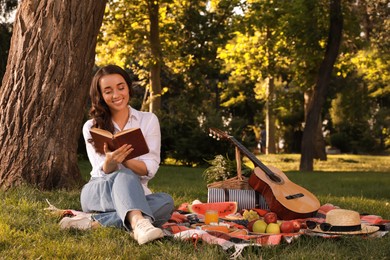 Photo of Happy young woman reading book on plaid in park. Summer picnic