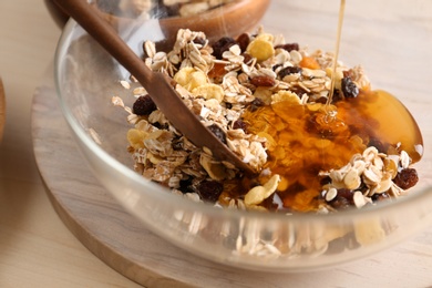 Photo of Pouring honey into bowl with oatmeal flakes and raisins on table, closeup. Healthy granola bar preparation