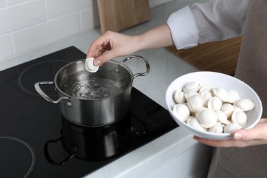 Photo of Woman putting frozen dumplings into saucepan with boiling water on cooktop in kitchen, closeup