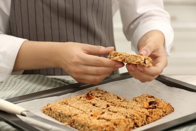 Photo of Woman taking granola bar from baking tray at table in kitchen, closeup