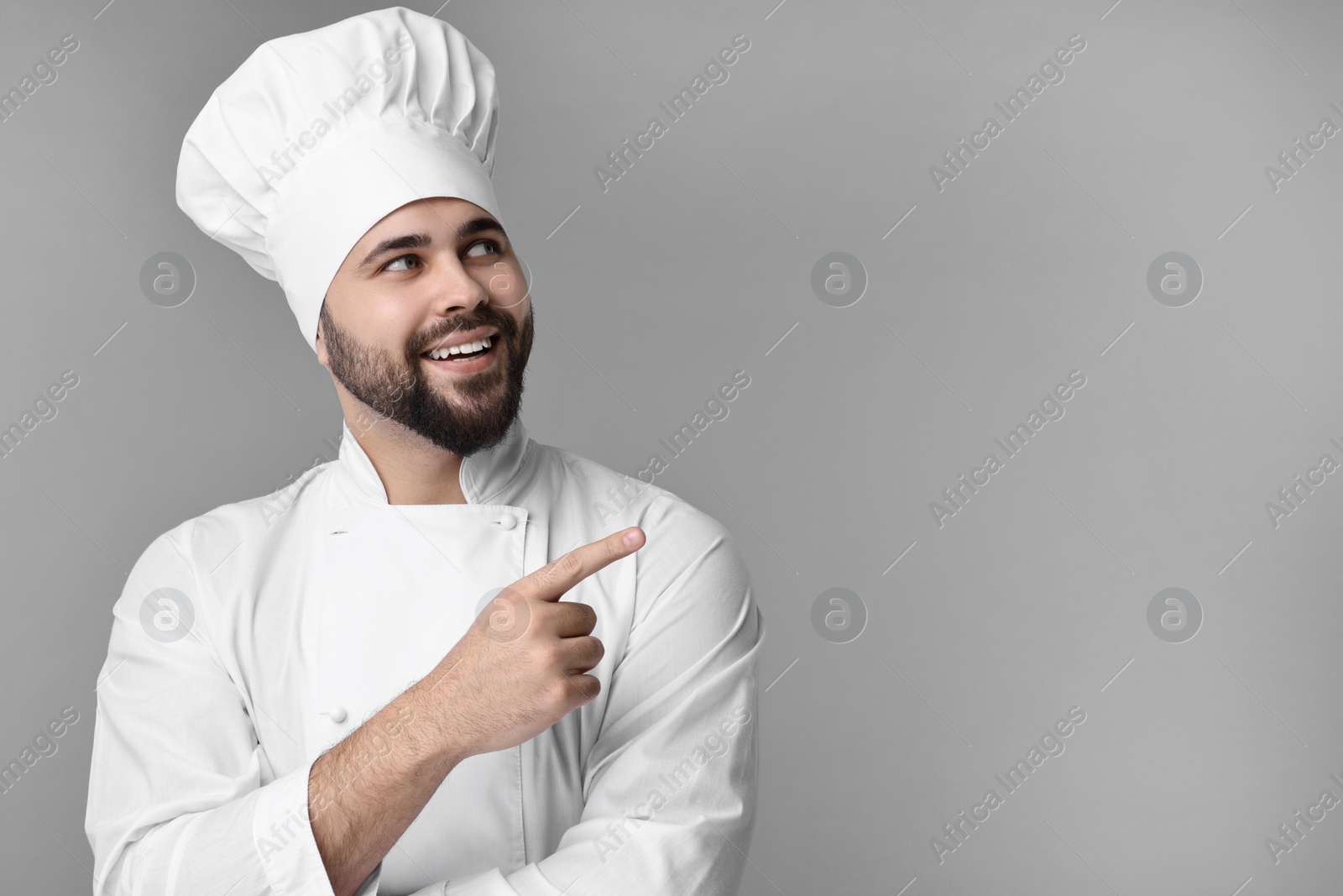 Photo of Happy young chef in uniform pointing at something on grey background. Space for text