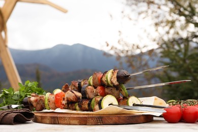 Metal skewers with delicious meat and vegetables served on wooden table against mountain landscape