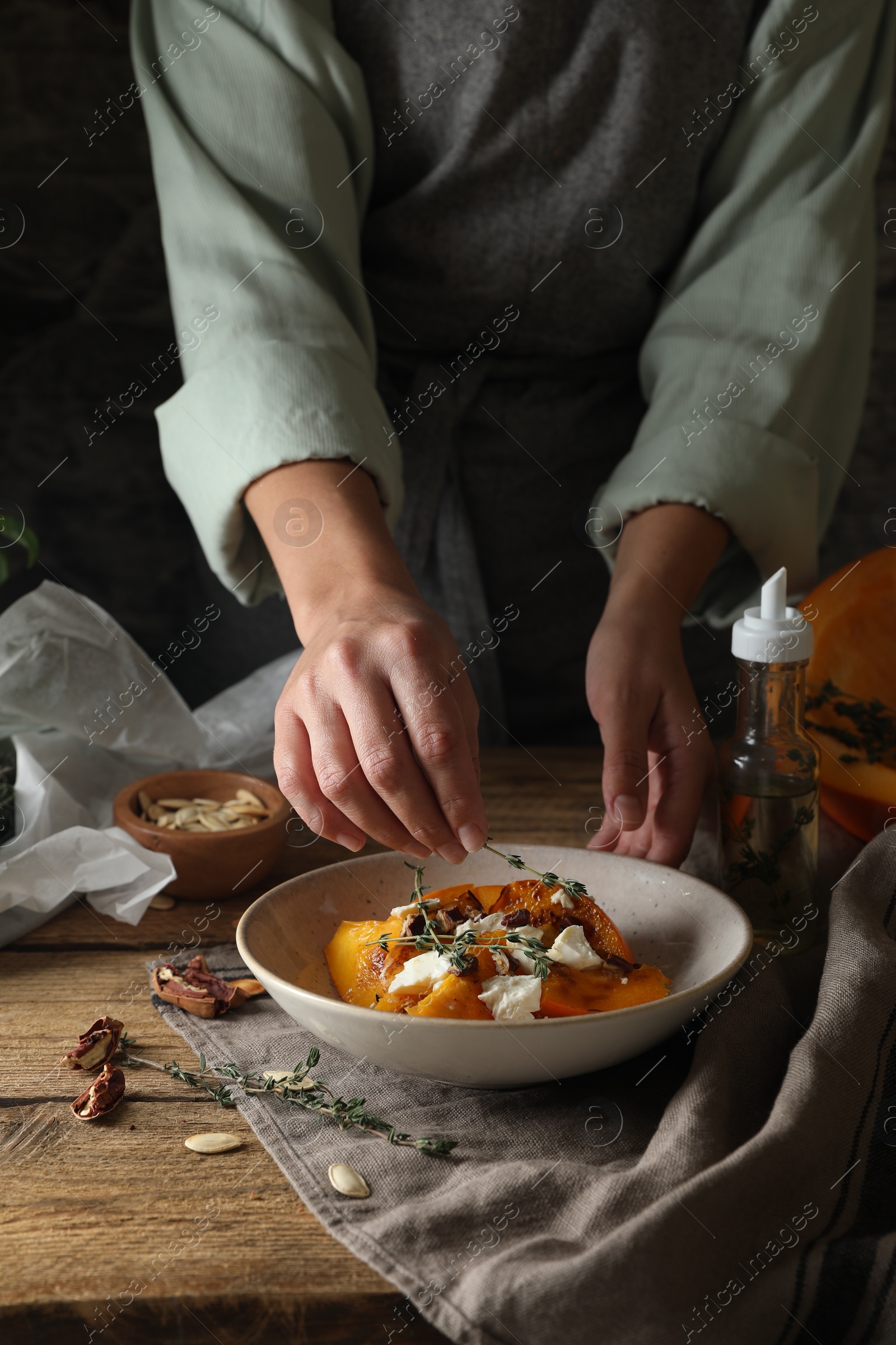 Photo of Woman adding aromatic thyme onto freshly baked pumpkin slices with cheese and pecans at wooden table, closeup