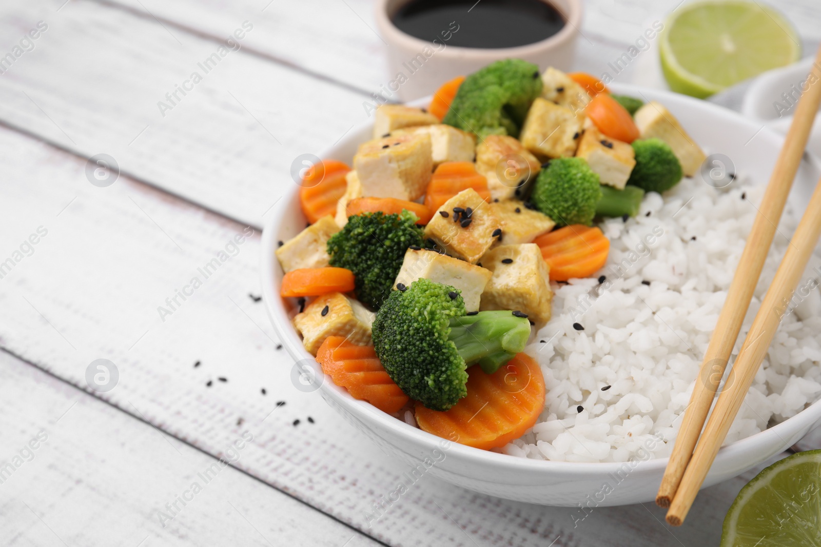 Photo of Bowl of rice with fried tofu, broccoli and carrots on white wooden table, closeup. Space for text