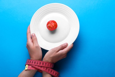 Woman holding plate with cherry tomato in hands tied with measuring tape on light blue background, top view. Diet concept