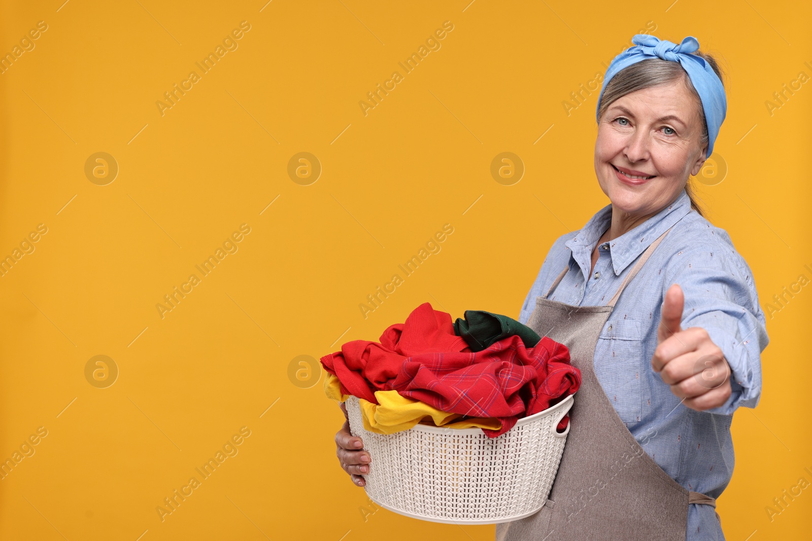 Photo of Happy housewife with basket full of laundry showing thumbs up on orange background, space for text
