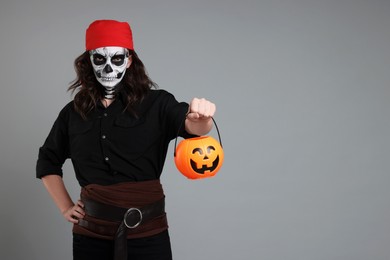 Photo of Man in scary pirate costume with skull makeup and pumpkin bucket on light grey background, space for text. Halloween celebration
