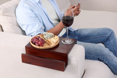 Glass of wine and snacks on sofa armrest wooden table. Man using smartphone at home, closeup