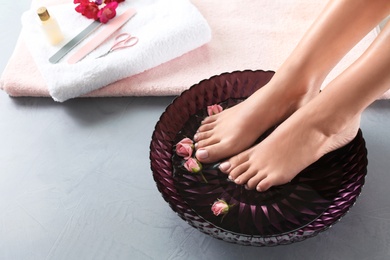 Closeup view of woman soaking her feet in dish with water and roses on grey floor, space for text. Spa treatment