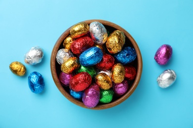 Wooden bowl with chocolate eggs wrapped in colorful foil on light blue background, flat lay