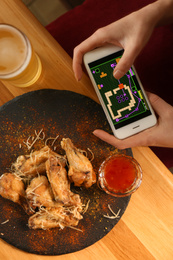 Photo of Woman playing game using smartphone at table with tasty BBQ wings, top view