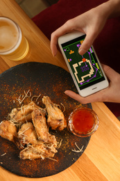 Woman playing game using smartphone at table with tasty BBQ wings, top view