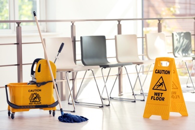 Photo of Safety sign with phrase Caution wet floor and mop bucket, indoors. Cleaning service