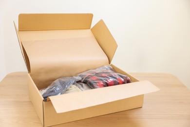 Photo of Open cardboard box with different clothes in plastic bags on wooden table. Delivery service