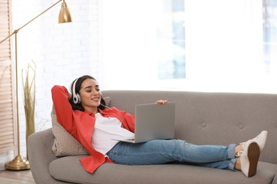 Photo of Young woman with headphones and laptop on sofa in living room