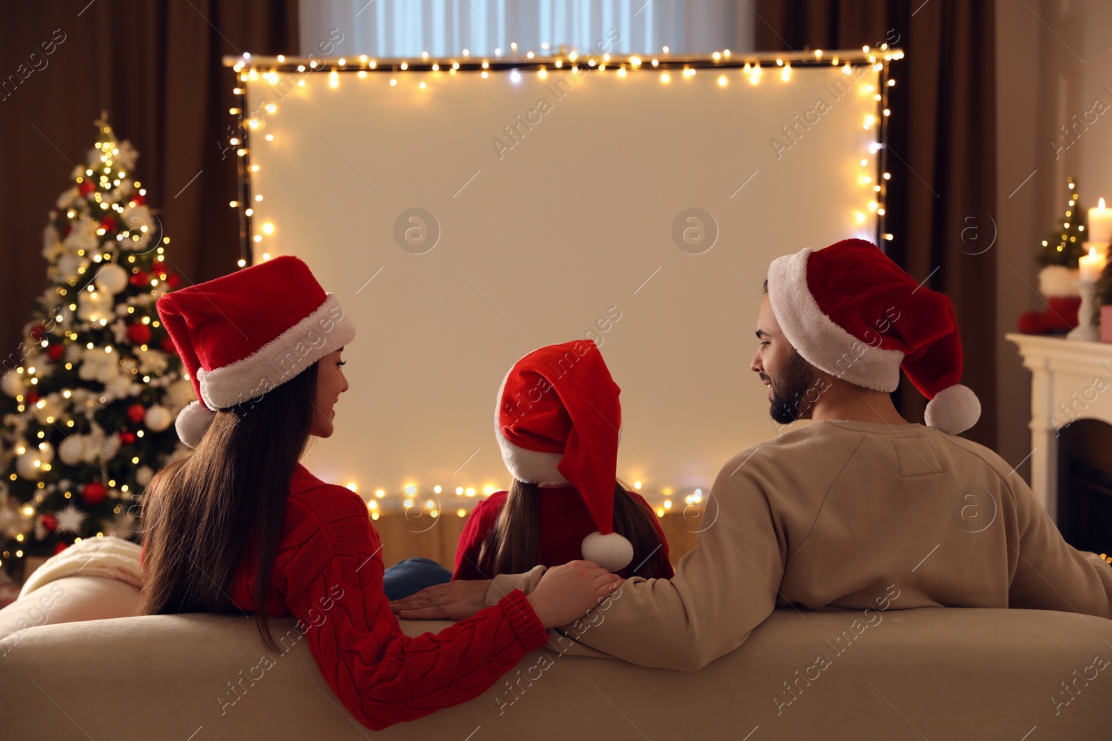 Photo of Family watching movie on projection screen in room decorated for Christmas, back view. Home TV equipment