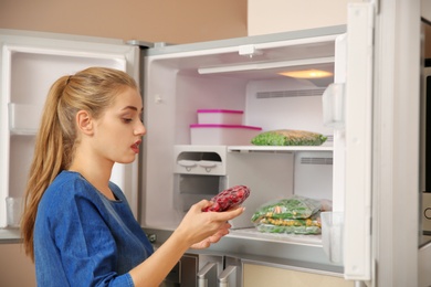 Photo of Young woman with frozen cherries near open refrigerator at home