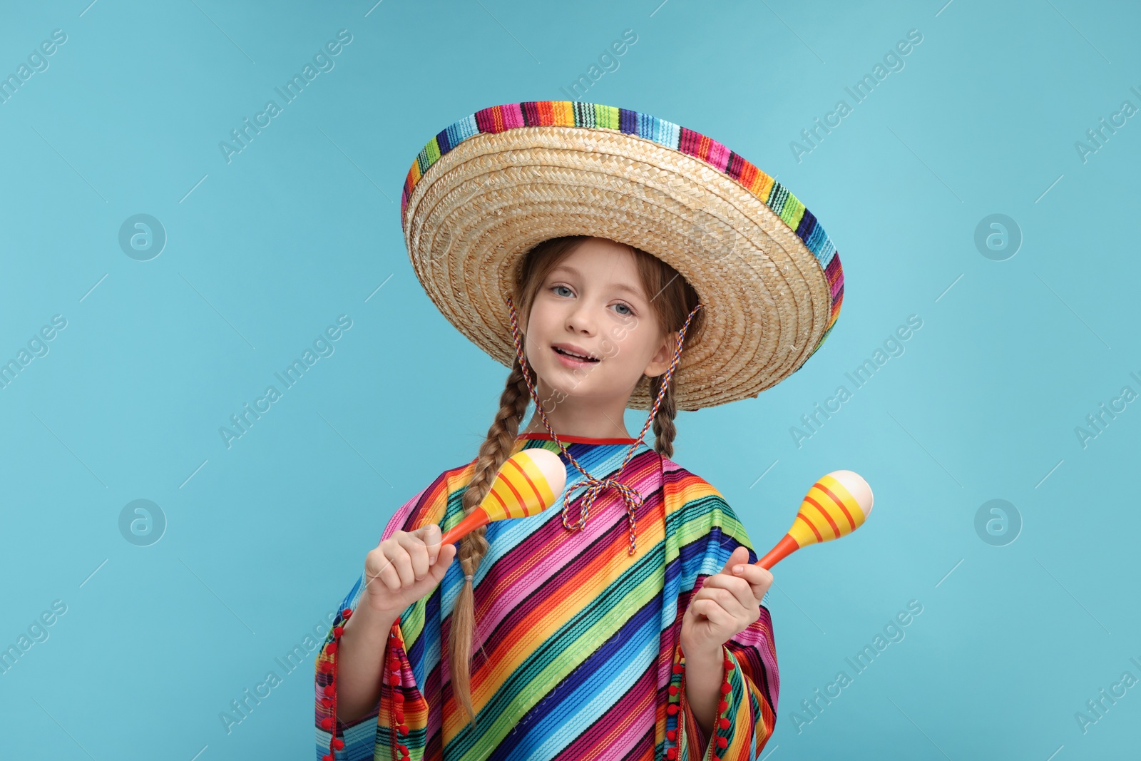 Photo of Cute girl in Mexican sombrero hat and poncho dancing with maracas on light blue background