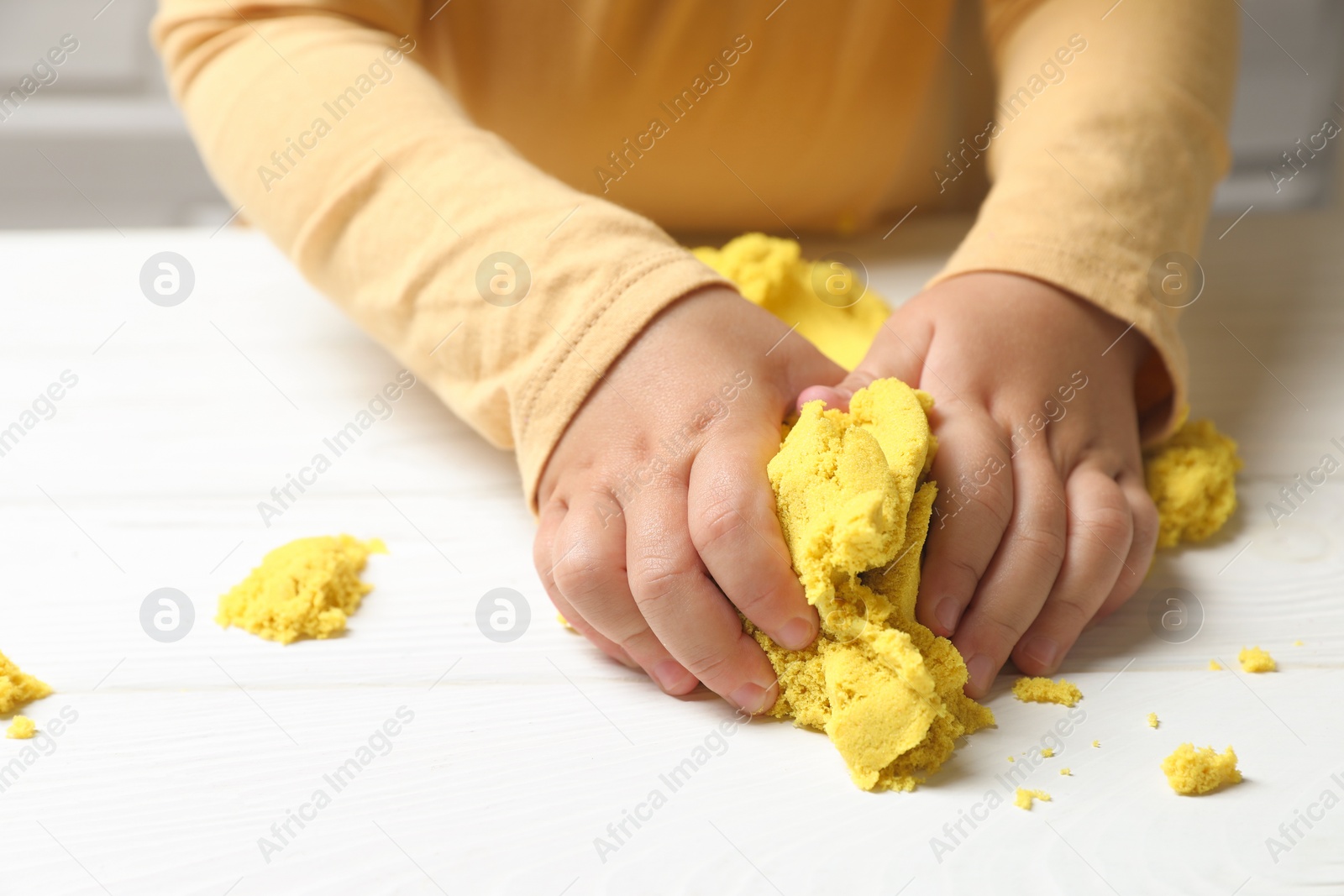 Photo of Little child playing with yellow kinetic sand at white wooden table, closeup