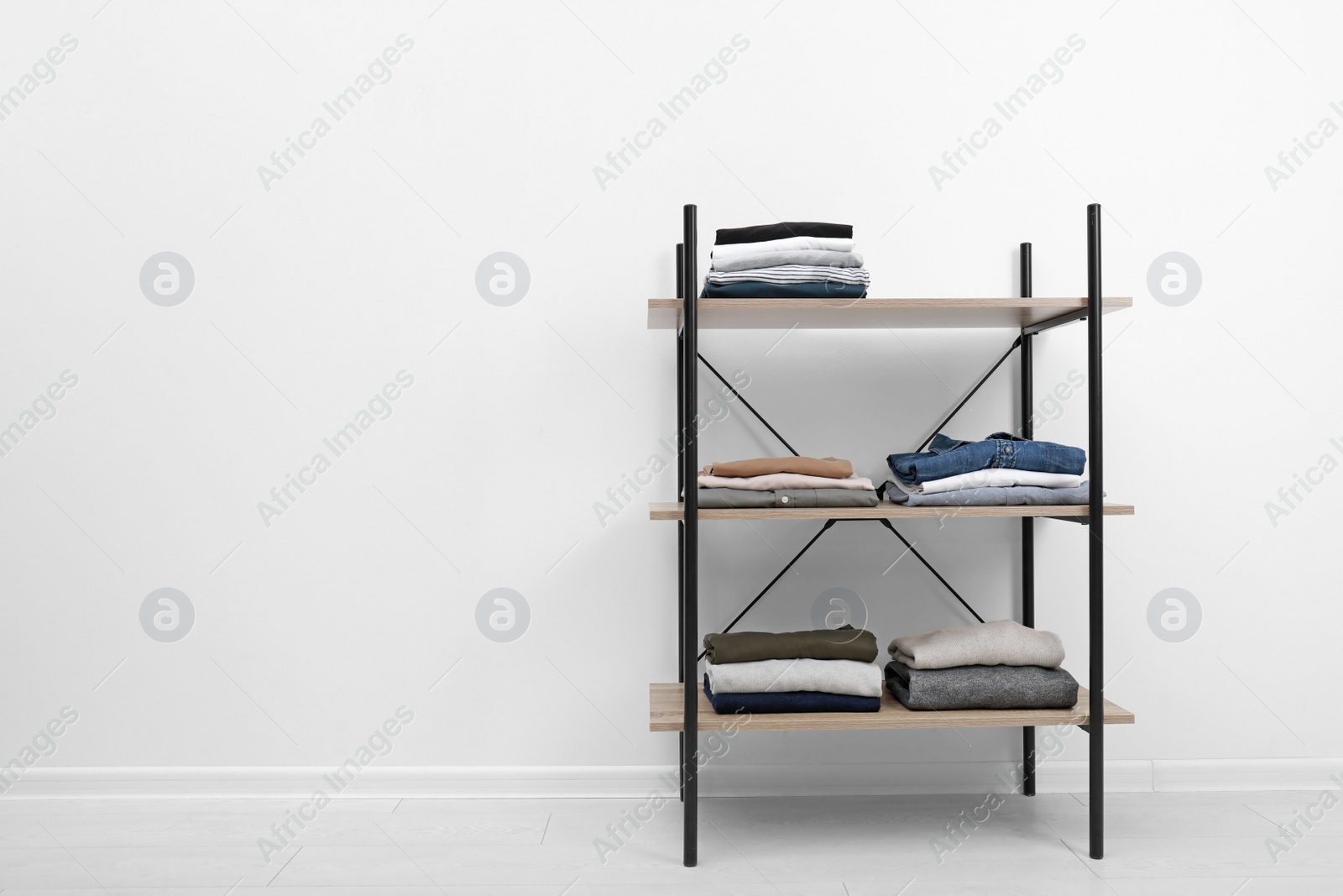 Photo of Modern shelves unit with stacked clothes on floor at white wall. Space for text
