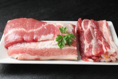 Photo of Pieces of raw pork belly and parsley on black textured table, closeup