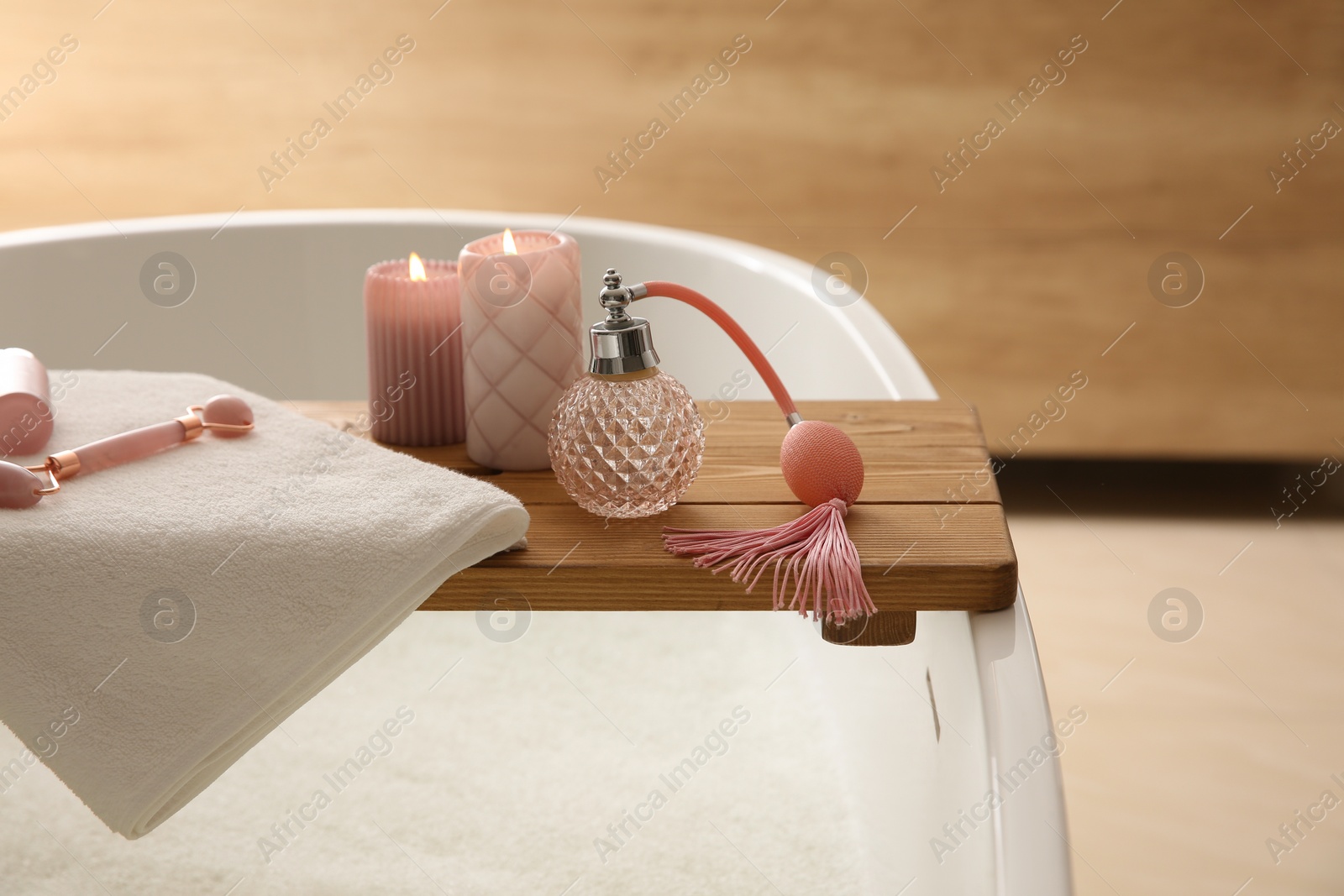 Photo of Wooden bath tray with face roller, cosmetic products and candles on tub indoors